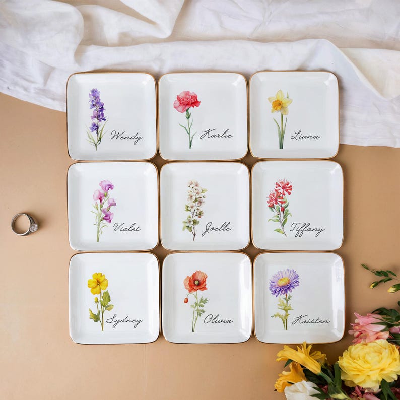 May include: Nine white ceramic ring dishes with gold trim, each featuring a different floral design and a name. The names are Wendy, Karlie, Liana, Violet, Joelle, Tiffany, Sydney, Olivia, and Kristen.