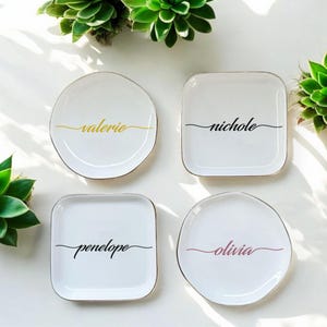 Op de afbeelding: Vier witte keramische ringenschalen met een gouden rand. Elke schaal heeft een andere naam in cursief erop geschreven: Valerie, Nichole, Penelope en Olivia. De namen zijn in zwart geschreven, behalve Olivia, dat in roze is geschreven.