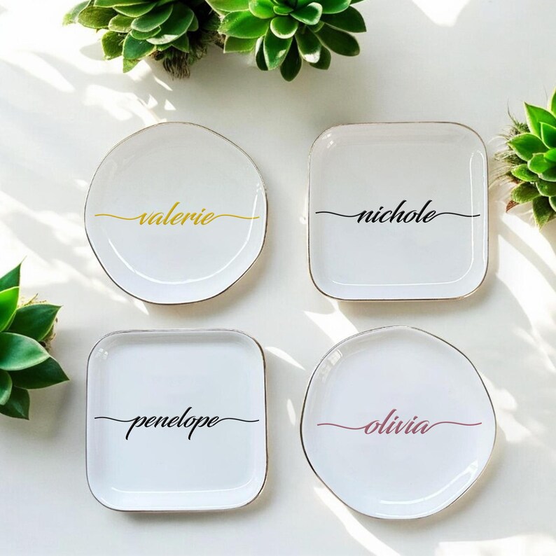 Op de afbeelding: Vier witte keramische ringenschalen met een gouden rand. Elke schaal heeft een andere naam in cursief erop geschreven: Valerie, Nichole, Penelope en Olivia. De namen zijn in zwart geschreven, behalve Olivia, dat in roze is geschreven.