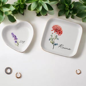 May include: Two ceramic trinket dishes with floral designs. The heart-shaped dish has a lavender design and the text "Lizzy". The square dish has a red chrysanthemum design and the text "Reina".
