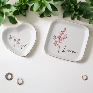 Puede incluir: Dos platos de cer&aacute;mica blancos para baratijas, uno en forma de coraz&oacute;n y otro cuadrado, con bordes dorados. El plato en forma de coraz&oacute;n tiene un dise&ntilde;o floral p&uacute;rpura y el nombre "Maria". El plato cuadrado presenta una rama floral rosa y el nombre "Lorena".