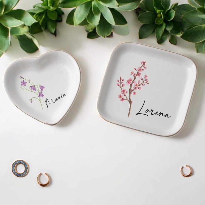 Puede incluir: Dos platos de cer&aacute;mica blancos para baratijas, uno en forma de coraz&oacute;n y otro cuadrado, con bordes dorados. El plato en forma de coraz&oacute;n tiene un dise&ntilde;o floral p&uacute;rpura y el nombre "Maria". El plato cuadrado presenta una rama floral rosa y el nombre "Lorena".