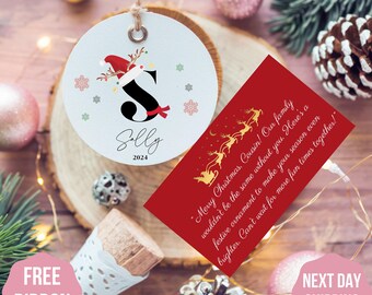 May include: Personalized Christmas ornament with a red and white Santa hat and the letter "S" in black. The ornament is decorated with snowflakes and the name "Sally" and the year "2024" are printed on the ornament. A red card with gold reindeer and the text "Merry Christmas. Cousin! Our family wouldn't be the same without you. Here's a festive ornament to make your season even brighter. Can't wait for more fun times together!" is also shown.