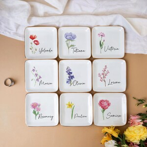 Puede incluir: Nueve platos cuadrados de cer&aacute;mica blanca con bordes dorados, cada uno con un dise&ntilde;o floral diferente y un nombre en escritura. Las flores incluyen amapolas, lavanda y rosas. Los nombres incluyen Yolanda, Tatiana y Victoria.