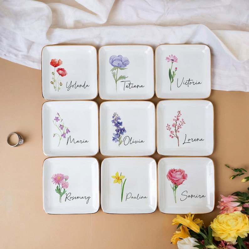 Puede incluir: Nueve platos cuadrados de cer&aacute;mica blanca con bordes dorados, cada uno con un dise&ntilde;o floral diferente y un nombre en escritura. Las flores incluyen amapolas, lavanda y rosas. Los nombres incluyen Yolanda, Tatiana y Victoria.