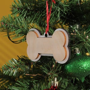 May include: A wooden dog bone-shaped Christmas ornament with a white border and a red ribbon for hanging. The ornament hangs on a green Christmas tree with green and red ornaments.