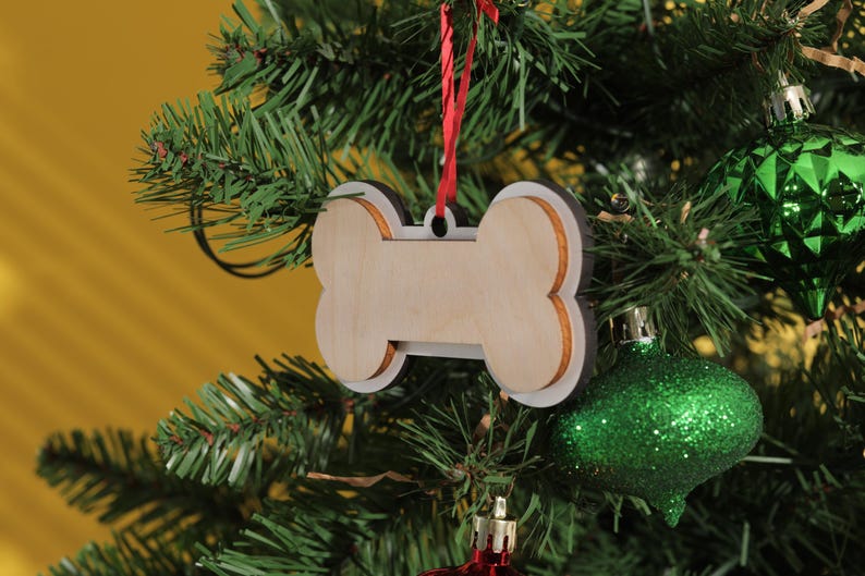 May include: A wooden dog bone-shaped Christmas ornament with a white border and a red ribbon for hanging. The ornament hangs on a green Christmas tree with green and red ornaments.