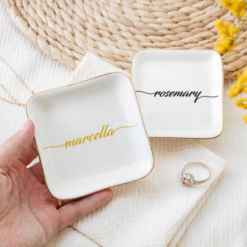 Op de afbeelding: Twee vierkante schalen van wit keramiek met een gouden rand. De schaal links heeft de naam "marcella" in gouden letters. De schaal rechts heeft de naam "rosemary" in zwarte letters.