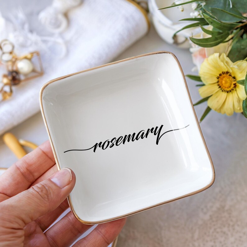 Op de afbeelding: Een wit keramiek sieradenbakje met een gouden rand. Het bakje heeft de tekst "rosemary" in zwarte, cursieve letters.