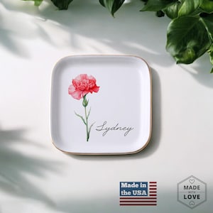 May include: White ceramic trinket tray with a gold rim. The tray features a watercolor illustration of a pink carnation and the name "Sydney".