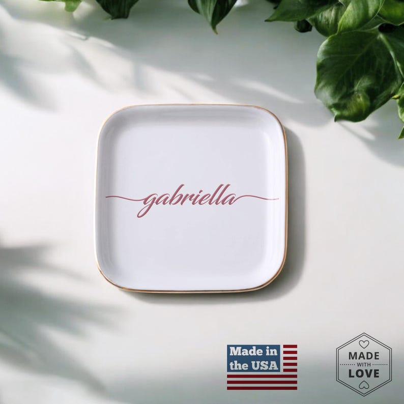 Op de afbeelding: Witte vierkante keramische sieradenbak met een gouden rand. De bak heeft de naam "gabriella" in roze cursief schrift.