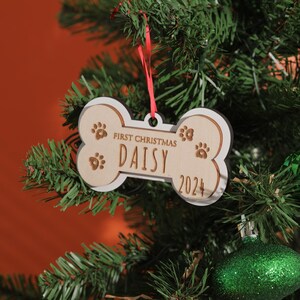 May include: A bone-shaped Christmas ornament with the text "First Christmas DAISY 2024" and paw prints. The ornament is made of light-colored wood with a white border and hangs from a red ribbon on a Christmas tree.