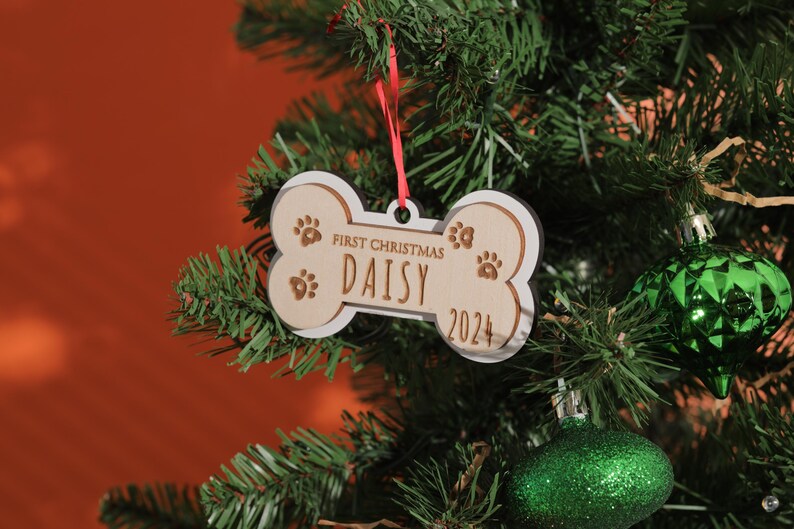 May include: A bone-shaped Christmas ornament with the text "First Christmas DAISY 2024" and paw prints. The ornament is made of light-colored wood with a white border and hangs from a red ribbon on a Christmas tree.