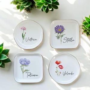 Puede incluir: Cuatro platos de cer&aacute;mica blanca con bordes dorados, cada uno con un dise&ntilde;o floral diferente y un nombre en escritura cursiva: Victoria, Stella, Tatiana y Yolanda. Las flores son de color rosa, morado y rojo.