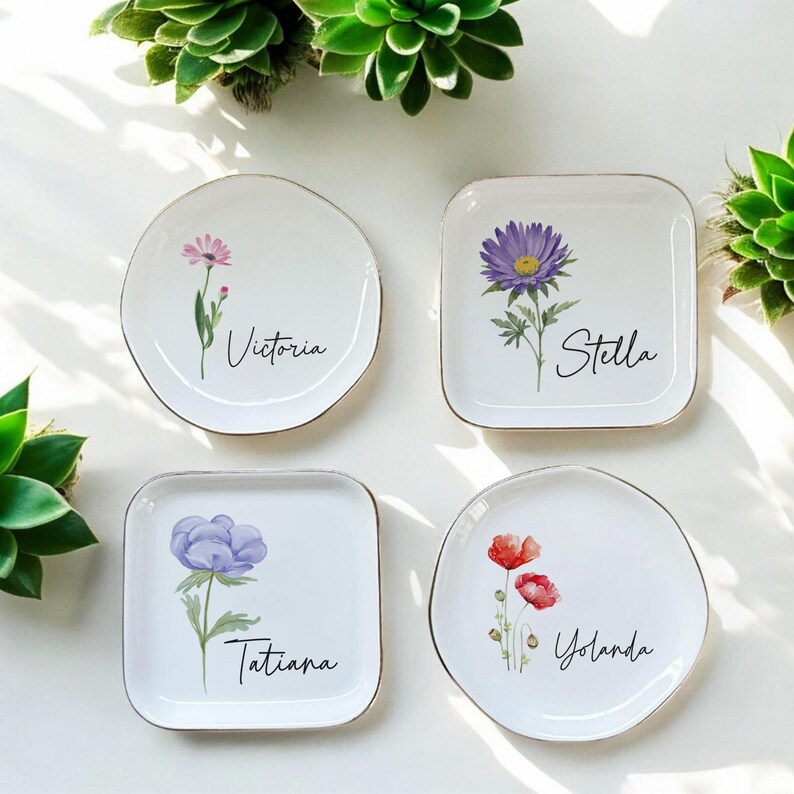 Puede incluir: Cuatro platos de cer&aacute;mica blanca con bordes dorados, cada uno con un dise&ntilde;o floral diferente y un nombre en escritura cursiva: Victoria, Stella, Tatiana y Yolanda. Las flores son de color rosa, morado y rojo.