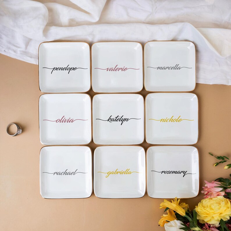 Op de afbeelding: Negen witte vierkante keramische ringschalen met een gouden rand en zwarte kalligrafie. Elke schaal heeft een andere naam erop geschreven: Penelope, Valerie, Marcella, Olivia, Katelyn, Nichole, Rachael, Gabriella en Rosemary.