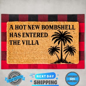 May include: A rectangular tan coir doormat layered over a red and black buffalo plaid mat. The doormat features the black text A HOT NEW BOMBSHELL HAS ENTERED THE VILLA and a black graphic illustration of three palm trees on the right side.