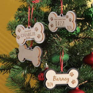 May include: Wooden dog bone-shaped Christmas ornaments hanging on a decorated tree. The ornaments are personalized with names and dates, including "Charlie," "Daisy," and "Barney." Each ornament has paw print details and is suspended by a red ribbon.