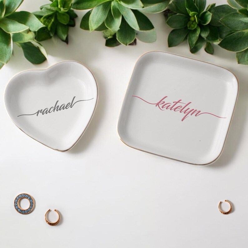 Op de afbeelding: Twee witte keramische sieradenbakjes met een gouden rand. Een bakje is hartvormig en heeft de tekst "rachael" in cursief. Het andere bakje is vierkant en heeft de tekst "katelyn" in cursief.