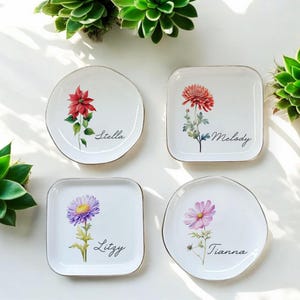 May include: Four white ceramic trinket dishes with gold rims, each featuring a different floral design and a name: Stella, Melody, Lizzy, and Tianna.