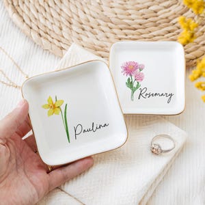 Puede incluir: Dos peque&ntilde;os platos cuadrados blancos con bordes dorados. Uno presenta un narciso amarillo y el nombre "Paulina". El otro tiene flores rosas y el nombre "Rosemary". Un anillo est&aacute; sobre una tela blanca.