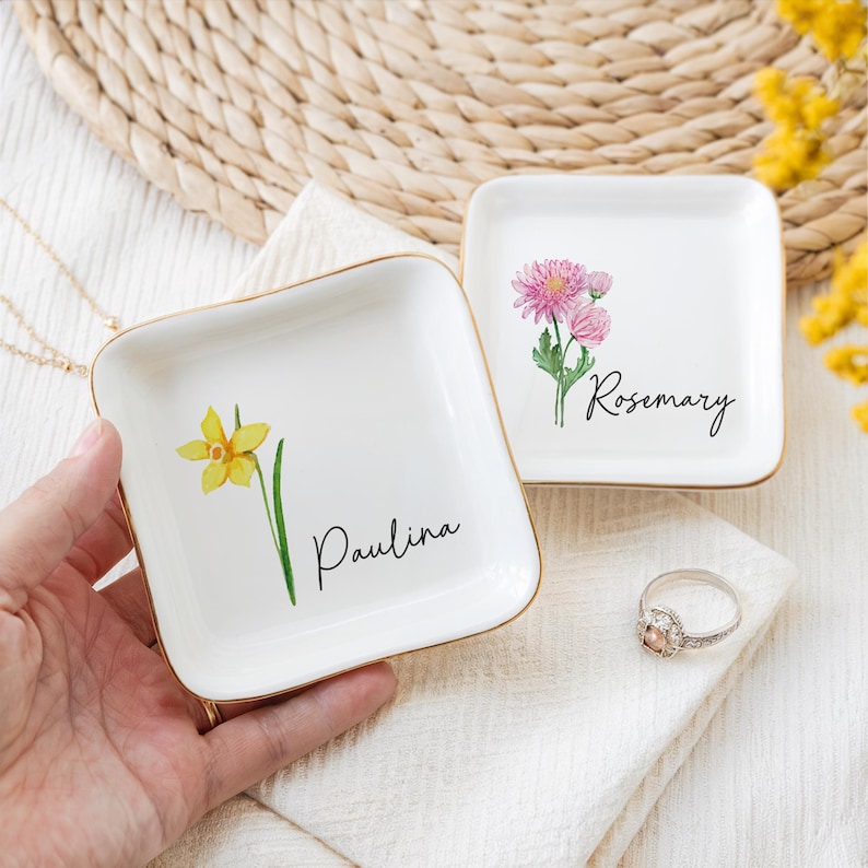 Puede incluir: Dos peque&ntilde;os platos cuadrados blancos con bordes dorados. Uno presenta un narciso amarillo y el nombre "Paulina". El otro tiene flores rosas y el nombre "Rosemary". Un anillo est&aacute; sobre una tela blanca.