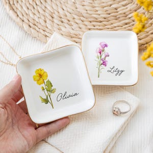 May include: Two white ceramic ring dishes with gold trim. The dish on the left features a yellow flower with green leaves and the name "Olivia". The dish on the right features a purple flower with green leaves and the name "Lizzy".