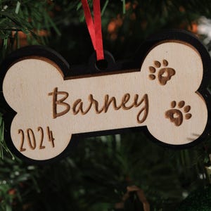 May include: A wooden dog bone ornament with the name "Barney" and the year "2024" engraved on it. The ornament has paw prints and a black border, hanging from a red ribbon. It is displayed on a Christmas tree.
