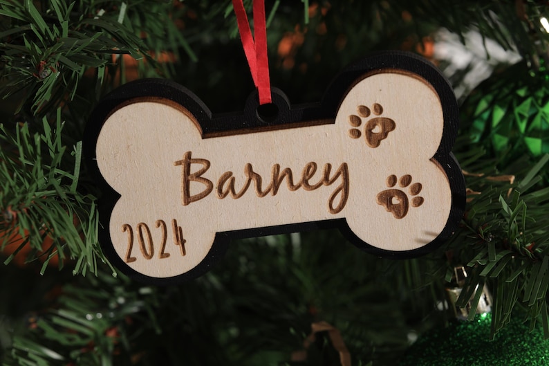 May include: A wooden dog bone ornament with the name "Barney" and the year "2024" engraved on it. The ornament has paw prints and a black border, hanging from a red ribbon. It is displayed on a Christmas tree.