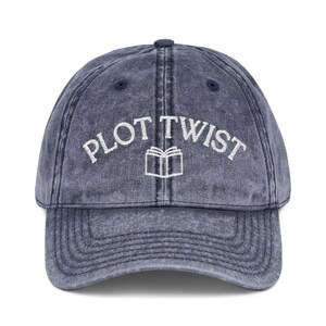 May include: A denim blue baseball cap with the words "PLOT TWIST" embroidered in white above a white book graphic. The hat has a curved brim and a distressed, vintage look.