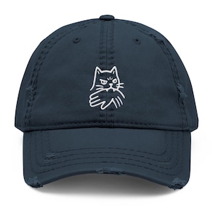 May include: Navy blue distressed baseball cap featuring a white embroidered cat design. The cat has an angry expression and is giving the middle finger. The cap has a curved brim and an adjustable strap.