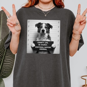 May include: Dark gray t-shirt featuring a black and white image of a dog in a mugshot pose. The dog holds a sign that reads "COUNTY JAIL ID: 042524 EMOTIONAL SUPPORT CRIMINAL". The shirt has a relaxed fit.