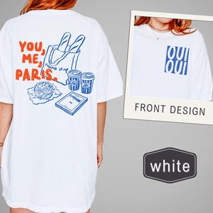 May include: White t-shirt with a blue and red graphic. The back design features the text "YOU, ME, PARIS" with illustrations of a baguette, coffee cups, and a pastry. The front design has the word "OUI" in blue.