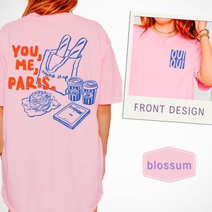 May include: Pink t-shirt with a graphic on the back featuring the words "YOU, ME, PARIS" in red and blue, along with illustrations of a baguette, coffee cups, and a pastry. A smaller image shows the front design with the text "OUI OUI".