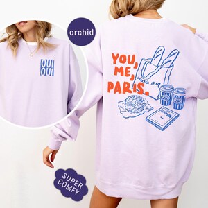 May include: Orchid-colored sweatshirt with the words "OUI OUI" in blue on the front. The back features a graphic with the words "YOU, ME, PARIS" in red, along with illustrations of a baguette, coffee cups, and a book.