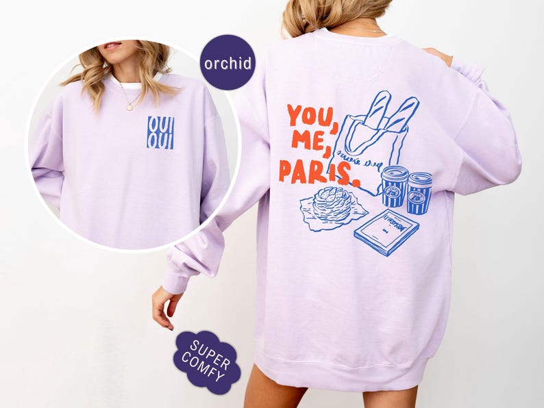 May include: Orchid-colored sweatshirt with the words "OUI OUI" in blue on the front. The back features a graphic with the words "YOU, ME, PARIS" in red, along with illustrations of a baguette, coffee cups, and a book.
