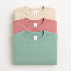 May include: Three crewneck sweatshirts in beige, rose, and mint green are stacked on top of each other. Each sweatshirt has a white tag at the neckline. The brand name "UNCOMMON CLOTH" is printed at the top of the image.