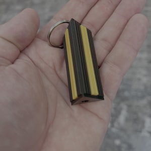 May include: A rectangular keychain with a silver ring. The keychain has a dark brown and yellow striped design. The keychain is held in a hand.