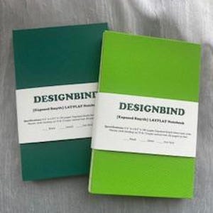 May include: Two notebooks, one dark green and one lime green, are displayed side-by-side. Each notebook is wrapped with a white band that reads "DESIGNBIND" in a bold, sans-serif font. The notebooks are approximately 5.75 x 8.25 inches.