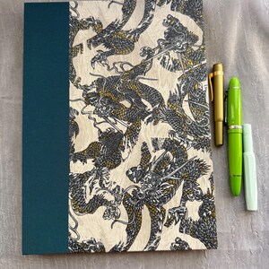 May include: A rectangular notebook with a solid dark teal spine and a cream-colored cover featuring a repeating pattern of gray and gold dragons. Three fountain pens are placed to the right: one matte gold, one bright lime green, and one pale mint green.