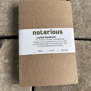 May include: A tan notebook with a white paper band featuring the text "notorious Layflat Notebook 5.5" w x 8.5" h (A5 size) LayflatNotebook 160 pages on 70 lb Cougar Natural text. Cover is 100% recycled handmade paper. Flush Cut, Exposed Spine.
