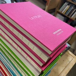 May include: A stack of colorful notebooks with pink, green, blue, and red covers. The top notebook is hot pink with the embossed text "LAYFLAT" and "DESIGNBIND" on the cover. The pages are white with visible stitching along the spine.