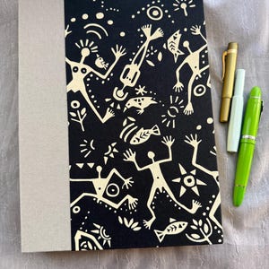 May include: A rectangular notebook with a beige fabric spine and a black cover featuring a cream-colored pattern of stylized human figures, fish, and sun symbols. Three fountain pens in gold, pale mint, and bright lime green lie beside it.