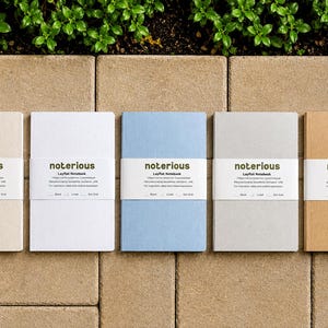 May include: Five rectangular notebooks in beige, white, light blue, gray, and tan, each featuring a white paper band with the text "notorious Layflat Notebook" and checkboxes for blank, lined, or dot grid page styles, arranged in a horizontal row.
