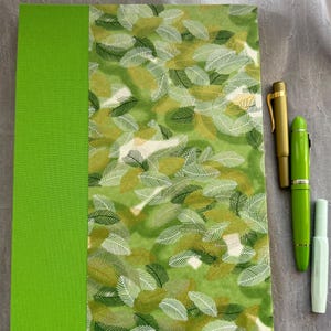 May include: A rectangular notebook with a solid lime green spine and a cover featuring a dense pattern of overlapping green and white leaves. Three pens are positioned to the right: one matte gold, one bright lime green, and one pale mint.