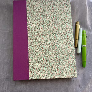 May include: A rectangular notebook featuring a solid purple spine and a cream-colored cover with a dense floral pattern of tiny red flowers and green leaves, accompanied by three fountain pens in gold, light blue, and bright green.