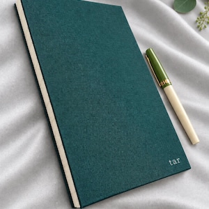 May include: A dark teal fabric-covered notebook with the text "tar" embossed in the bottom right corner, featuring cream-colored pages. Beside the notebook lies a fountain pen with an olive green cap and a cream-colored barrel with gold accents.