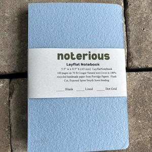 May include: A light blue layflat notebook with a textured cover and a white paper band featuring the text: notorious Layflat Notebook 5.5" w x 8.5" h (A5 size) 160 pages on 70 lb Cougar Natural text. Cover is 100% recycled handmade paper.