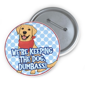 Op de afbeelding: Een ronde button met een cartoonillustratie van een lachende gele Labrador met een rode bandana. De button heeft de tekst "WE'RE KEEPING THE DOG, DUMBASS!" op een blauwe stippelachtergrond, met een rode rand en een zilveren speld aan de achterkant.