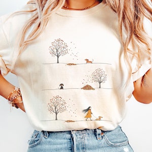 May include: A woman wearing a light beige t-shirt with a graphic of three trees with falling leaves and a dachshund walking in each scene. The text "Ivory" is visible at the bottom of the image.